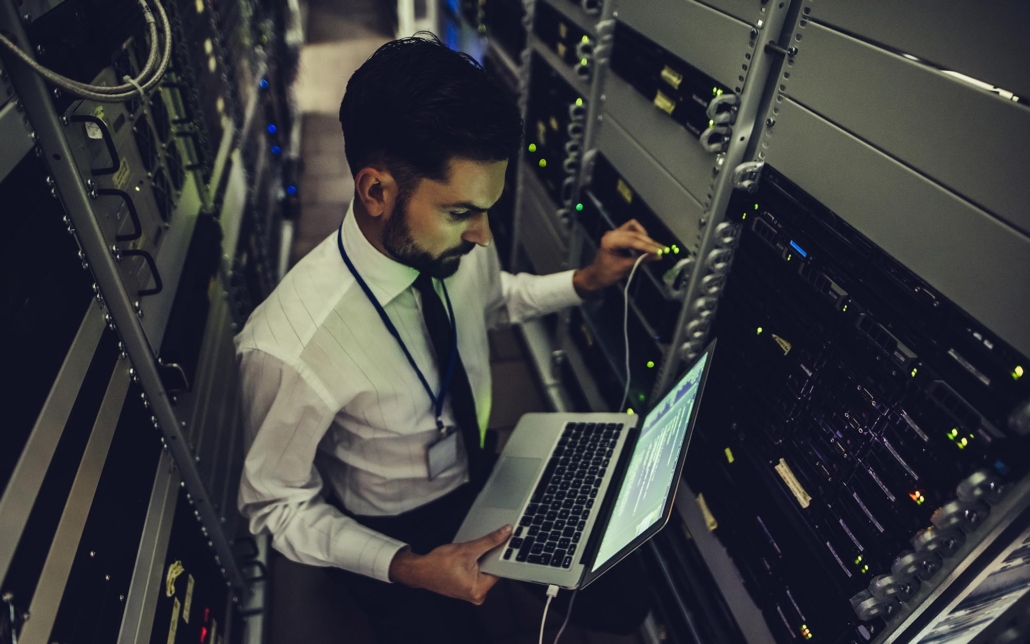 Image of a business person in a server room, looking at a laptop.